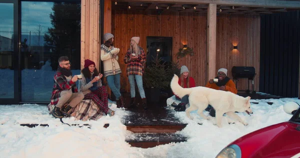 Kelompok bahagia santai multietnis teman minum teh panas di nyaman rumah musim dingin teras dengan anjing, mobil merah gerakan lambat Stok Foto