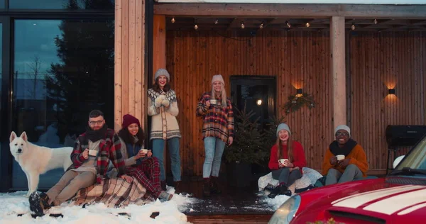 Mooie kerstvakantie. Groep van gelukkige vrienden met warme dranken kijken naar de camera op het terras van het winterhuis slow motion. Stockafbeelding