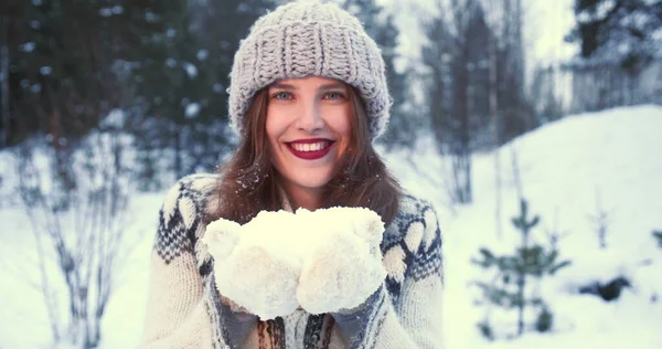 Winnaarsmod. close-up mooi jong gelukkig vrouw in hoed, wanten blaast op sneeuw in handen op camera bij bos slow motion Stockfoto