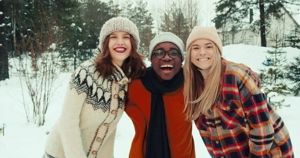 Wintervakantie samen. Drie multi-etnische vrolijke vrienden zwaaien, glimlachen naar de camera op mooie besneeuwde bos slow motion. Rechtenvrije Stockfoto's