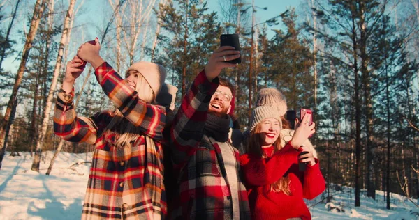 SELFISK KULTUR. Kamera bevæger sig rundt glade multietniske venner tager selfies på solrige snedækket vinter skov slow motion. Royaltyfrie stock-billeder