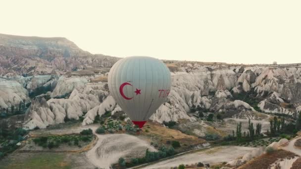 JUL 26, 2021 CAPPADOCIA Tiro aéreo de grande balão de ar quente branco com símbolo da Turquia voando acima das rochas no céu do nascer do sol. — Vídeo de Stock