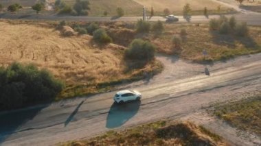 Yüksek açılı insansız hava aracı takibi, beyaz araba güneşli arazide güzel bir yolda ilerliyor, yolda birleşiyor..