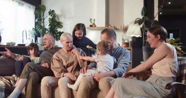 MULTI GENERATIONAL FAMILY Vrolijke senior grootouders, jonge mama en papa, tieners en baby meisje praten op de bank thuis. — Stockvideo