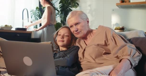 Amistad familiar y vínculo. Feliz abuelita de 70 años de edad usando el ordenador portátil hablando con una linda adolescente en el acogedor sofá del hogar. — Vídeo de stock