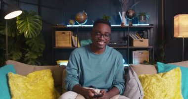 Genç mutlu gen- z siyah öğrenci başını sallıyor, müzik eşliğinde dans ediyor evdeki kanepede akıllı telefon uygulaması kullanıyor..