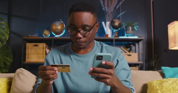ONLINE FIZETÉSEK Boldog fókuszált fekete üzletember olvas, belép a hitelkártya adatait okostelefon bevásárló app otthon. — Stock videók