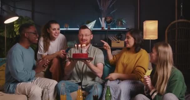 BIRTHDAY CAKE Feliz sorrindo homem de cabelos vermelhos sopra no bolo na festa de confete divertido comemorando com amigos multiétnicos. — Vídeo de Stock