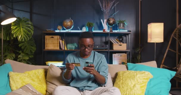Compras concepto en línea. Feliz hombre negro empresario introduce los detalles de la tarjeta de crédito utilizando la aplicación del teléfono inteligente en el sofá acogedor. — Vídeos de Stock