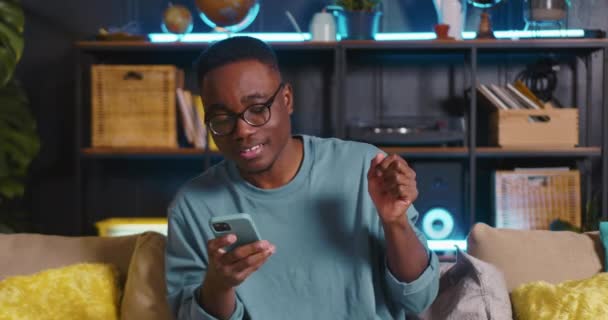 Portrét šťastný vzrušený africký muž blogger přikyvující hlavu k hudbě nakupování on-line pomocí aplikace chytrého telefonu v útulném domě. — Stock video