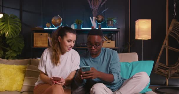 Glad ung svart man och vit kvinna använder mobiltelefoner surfa internet tillsammans, spela spel online hemma. — Stockvideo