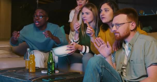 Felices jóvenes fanáticos de los deportes multiétnicos ven el partido de fútbol en la televisión en casa con aperitivos y bebidas, celebran el gol y ganan. — Vídeo de stock