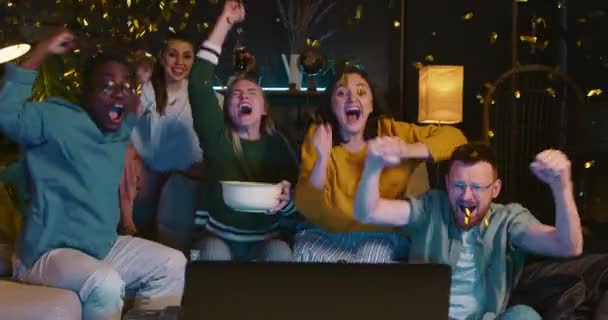 Fani sportu świętują bramkę. Podekscytowani młodzi wielonarodowi przyjaciele oglądają mecz w telewizji w domu wspierając zespół konfetti. — Wideo stockowe