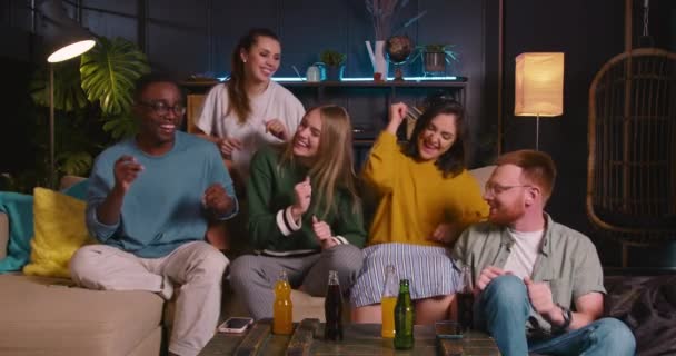 Grupo de divertidos jóvenes multiétnicos felices bailan juntos sentados en el sofá celebrando el éxito del logro ganador. — Vídeo de stock