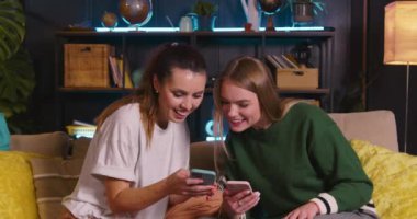 İki mutlu genç Avrupalı güzel arkadaş akıllı telefonlar kullanarak online alışveriş yapıyorlar, rahat oturma odasında kanepede gülüyorlar..
