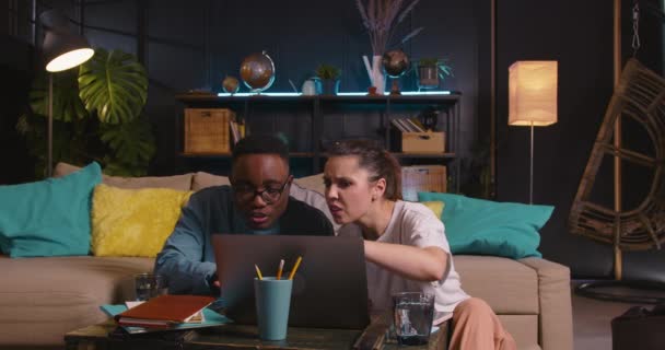 Två glada unga kreativa multietniska studenter vänner arbeta tillsammans med hjälp av laptop sitter på golvet ler mot mysiga hem. — Stockvideo