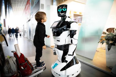Siyah tulumlu küçük bir çocuk valiz için tramvayda duruyor ve halka açık bir yerde duran ve gülümseyen bir robota ilgiyle bakıyor..