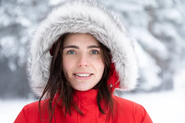 Portrait of a beautiful young smiling brunette with gray eyes in a red jacket with a hood made of faux fur on a winter day against the background of snowy trees. — Stock Photo, Image