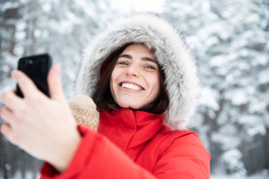 Güzel, genç, gülen esmer, gri gözlü, kapüşonlu, sahte kürklü bir kadının portresi. Kış günü, arka plandaki karlı ağaçlara karşı. Cep telefonuyla selfie çeken ve konuşan bir kadın.