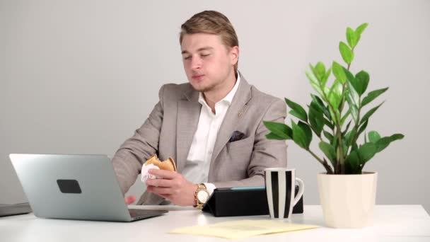 Filmación del joven sentado en el escritorio del trabajo y comiendo hamburguesa, hora del almuerzo — Vídeos de Stock
