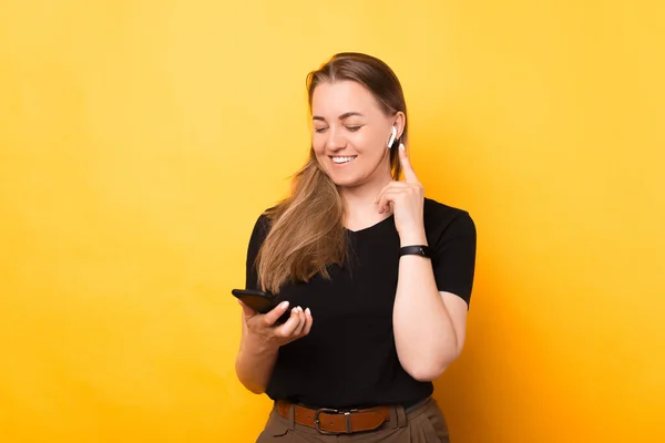 Beautiful woman is connecting to phone her new wireless ear phones. Portrait over yellow background