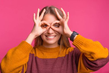 Genç oyuncu kadın kameraya gülümserken pembe arka planda dürbün hareketi yapıyor..