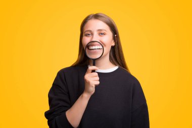 Gülümseyen genç kız gülümsemesinin üzerinde bir büyüteç tutuyor. Sarı arkaplanda fotoğraf.