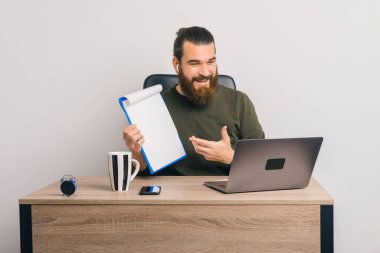 Ofiste oturan sakallı hippi bir video görüşmesi yapıyor. Kağıt panodan bazı veriler sunuyor..