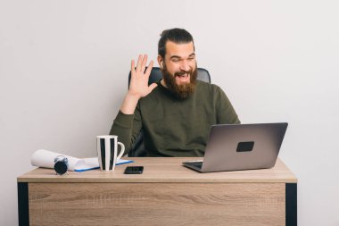 Genç sakallı adam ofiste oturuyor, konuşuyor ve web kamerasında el sallıyor, dizüstü bilgisayarı masanın üstünde..
