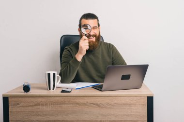 Neşeli sakallı erkek işçi, büyüteçle kameraya bakarken ofiste oturuyor..