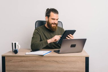 Ofiste çalışan gülümseyen sakallı adam masadaki bilgisayarı açarken tabletini kullanıyor..