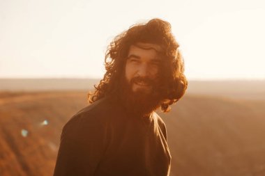 Gün batımında tepede rüzgarlı bir günde uzun saçlı sakallı hippi portresi.