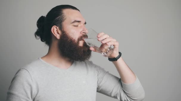 Bärtiger Mann steht vor grauem Hintergrund und trinkt ein Glas Wasser — Stockvideo