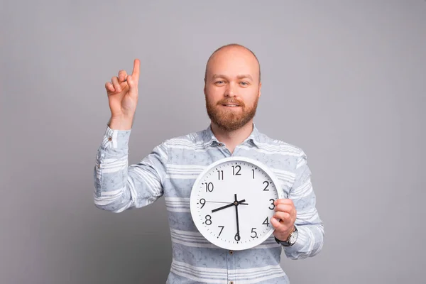 Joven hombre barbudo guapo sosteniendo el reloj y apuntando hacia arriba en el espacio de copia — Foto de Stock