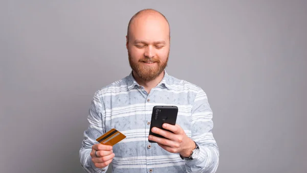 Hombre barbudo alegre en pago casual con tarjeta de crédito y teléfono inteligente — Foto de Stock