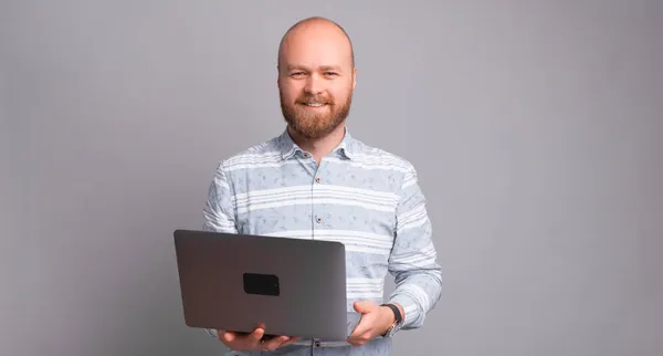 Hombre feliz con barba sosteniendo portátil sobre fondo gris y mirando a la cámara — Foto de Stock