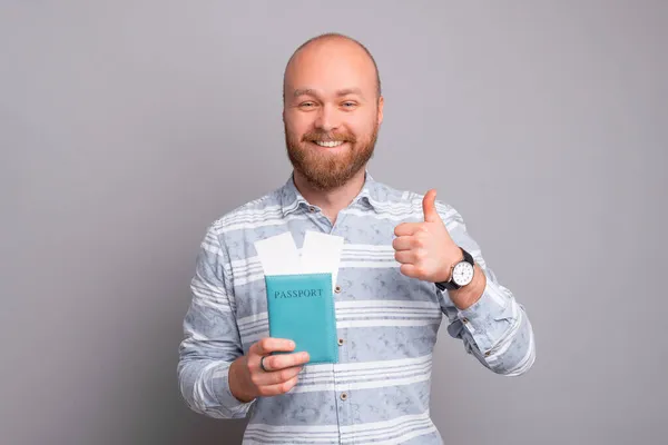 Joven sonriente con barba sosteniendo pasaporte y billetes de avión y mostrando el pulgar hacia arriba — Foto de Stock