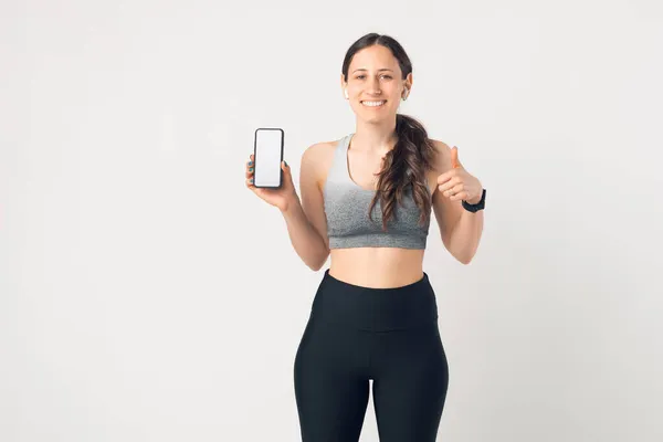Ευτυχισμένη νεαρή αθλητική γυναίκα που δείχνει την οθόνη του τηλεφώνου και καθιστώντας τον αντίχειρα μέχρι χειρονομία πάνω από λευκό τοίχο — Φωτογραφία Αρχείου