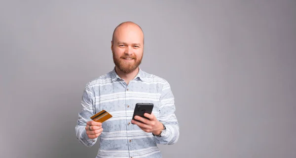 Hombre barbudo guapo en casual usando tarjeta de débito y teléfono inteligente fro pagos en línea sobre fondo gris — Foto de Stock