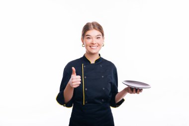 İzole edilmiş beyaz arka planda aşçı üniforması giyen genç bir kadın. Bir tabak fırlatmak ve baş parmaklarınla iyi bir jest yapmak, mutlu ve neşeli gülümsemek.