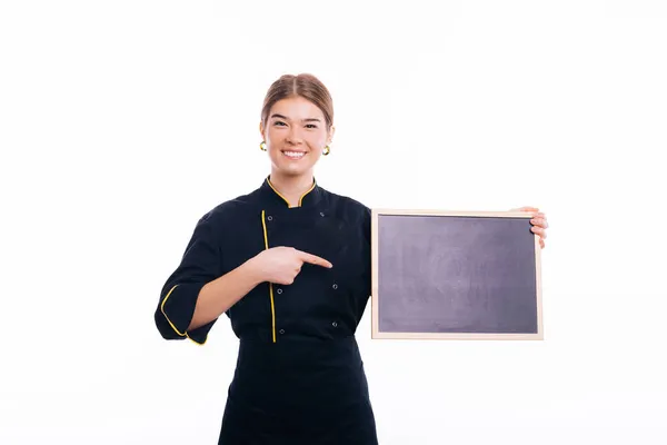 Veselá mladá žena, kuchařka v uniformě, drží tabuli a ukazuje na ni — Stock fotografie