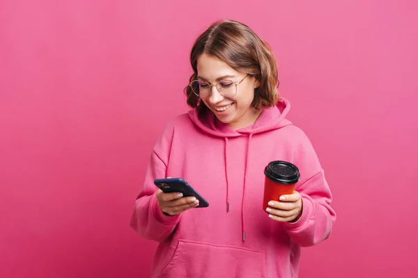 Jovem feliz com capuz rosa usando smartphone e segurando xícara de café — Fotografia de Stock