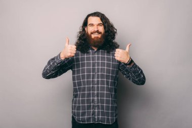 Kareli gömlekli mutlu sakallı genç adam gri arka planda baş parmak gösteriyor.