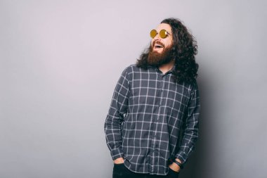 Gülen sakallı hippi adamın uzun saçlı ve güneş gözlüklü fotoğrafı gri arka plana bakıyor..