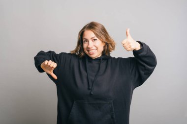 İster beğen ister beğenme, genç, kendine güvenen bir kadın parmak kaldırıyor ve gri arka planda baş parmağını öne eğiyor.