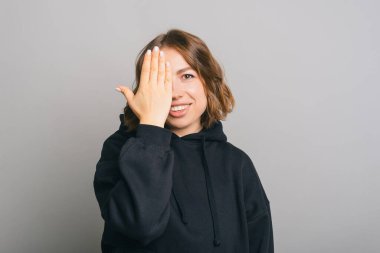 Siyah kapüşonlu genç ve güzel bir kadın gülümsüyor ve gri arka planda avuç içi ile yüzünü kapatıyor.