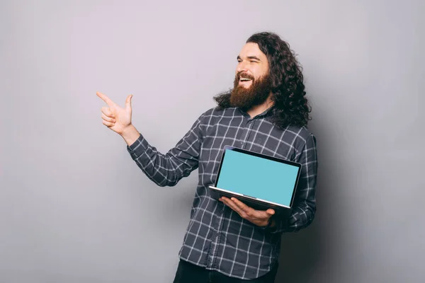 Portrait de jeune homme barbu pointant du doigt sur fond gris et tenant un ordinateur portable avec écran blanc — Photo