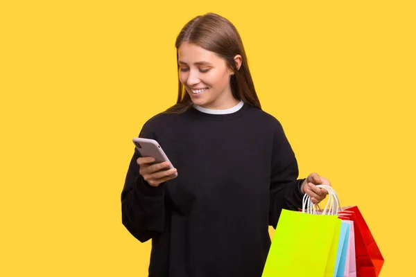 Szczęśliwa uśmiechnięta kobieta z torbami na zakupy patrzy na swój telefon. — Zdjęcie stockowe