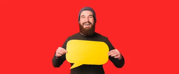 Banner μέγεθος φωτογραφία του χαμογελαστού νεαρός άνδρας με γενειάδα κρατώντας κενό κίτρινο φούσκα ομιλία — Φωτογραφία Αρχείου