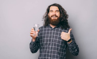 Kareli tişörtlü, elinde bir bardak su tutan ve baş parmağını gösteren gülümseyen genç adamın portresi.